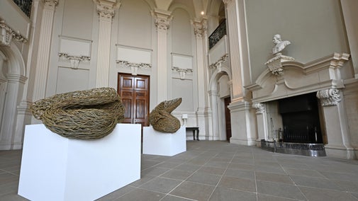 Two willow sculptures on white plinths in a large historic stone hall with carved architecture